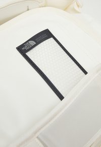Sac blanc avec une poche en mesh noir arborant le logo "The North Face". Détails en tissu texturé et en couture à l'extérieur.