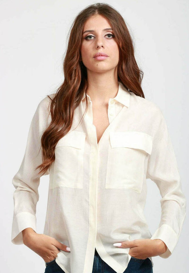 Blusa crema leggera e semi-trasparente con chiusura a bottoni frontale, due tasche sul petto e maniche lunghe. Presenta un colletto classico e una vestibilità morbida.