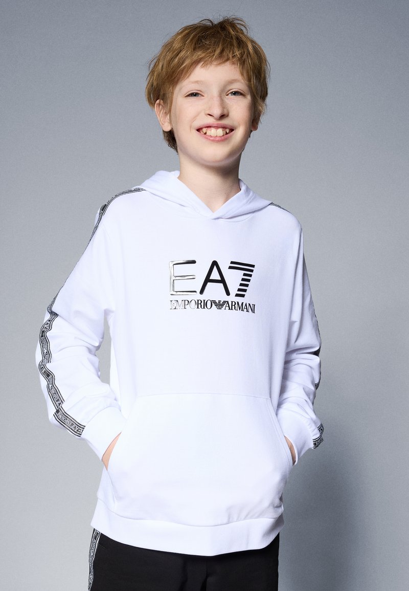 Fehér kapucnis pulcsi elülső zsebbel, ezüstszínű "EA7 Emporio Armani" logóval és mintás fekete díszítésekkel az ujjakon. Az anyag puhanak tűnik.
