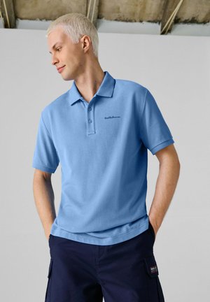 Giovane uomo con capelli biondi corti che indossa una polo azzurro chiaro della Red Bull Racing e pantaloni blu scuro, in piedi con le mani nelle tasche.