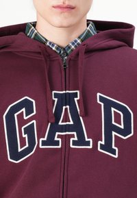 Sudadera con cremallera color granate con un gran gráfico de "GAP" en azul marino y blanco. Cuenta con cordones y una textura de forro polar, combinada con una camisa de cuadros.