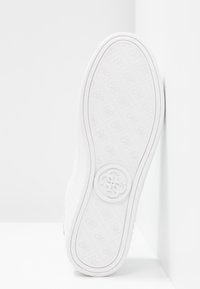 Semelle de basket blanche avec une adhérence texturée, présentant un logo circulaire au centre. Le bord est subtilement incurvé, mettant en valeur un design épuré.