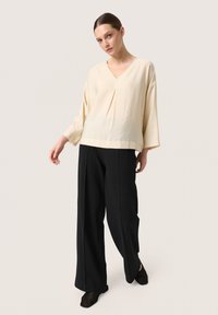 Blusa beige chiaro con scollo a V e maniche a tre quarti, abbinata a pantaloni neri a gamba larga con vita alta e scarpe nere slip-on.