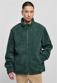 Giacca in pile verde con colletto alto e zip completa, caratterizzata da un corpo texturizzato e tessuto morbido, abbinata a pantaloni color sabbia.