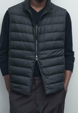 Homme portant une veste noire matelassée sans manches zippée sur une chemise noire à manches longues, les mains dans les poches d'un pantalon foncé, sur un fond clair uni.