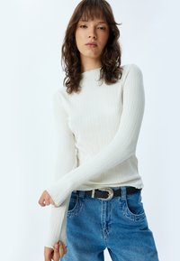 Maglietta bianca a maniche lunghe con collo alto; tessuto a righe; abbinata a jeans in denim blu e una cintura nera con fibbia metallica.