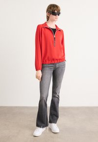 Rode pullover met een kraag en ritssluiting, gecombineerd met grijze flare jeans en witte sneakers. Model draagt een zonnebril en staat op een glad oppervlak.
