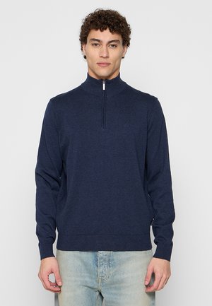 Mann steht frontal, trägt einen dunkelblauen Zip-Up-Pullover und hellblaue Jeans vor einem schlichten weißen Hintergrund.