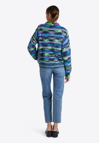 Ce pull coloré en tricot présente des rayures verticales en bleu, vert et violet, avec un col montant et une coupe décontractée. Associé à un jean bleu.