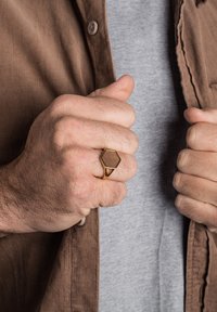 Gold metal ring with a hexagonal wooden inset. Hand is gripping a brown corduroy shirt, worn over a gray t-shirt. Textured details visible.