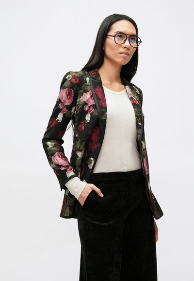 Blazer floral avec un fond noir, présentant des roses roses, rouges et vertes. Associé à un haut gris clair et un pantalon texturé noir.