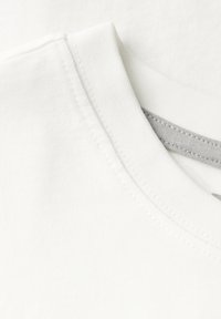 Bílý tričko s kulatým výstřihem, hladkým bavlněným povrchem a subtilními švy. Na obrázku jsou viditelné vrstvy látky.