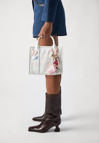 Borsa in pelle bianca con il testo "LIFE IS A FAIRYTA", decorata con charm colorati e fiocchi. Presenta manici corti e una forma strutturata.
