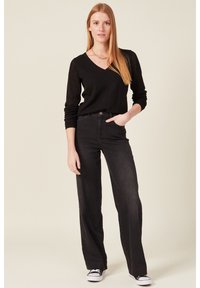 Schwarzer V-Ausschnitt-Pully mit strukturierten Ärmeln, kombiniert mit hochgeschnittenen schwarzen Jeans. Das Model trägt schwarze Sneakers und minimalistischen Schmuck vor einem neutralen Hintergrund.