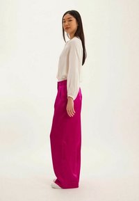 Pantalons larges fuchsia à texture lisse, taille haute et plis, associés à un haut blanc à manches longues dans une coupe décontractée.