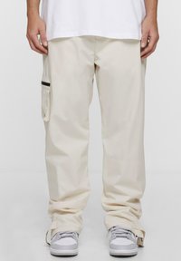 Pantaloni cargo beige con una vestibilità rilassata, dotati di tasca laterale, dettagli neri e zip alla caviglia. Abbinati a sneakers grigie.