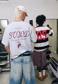 Grå oversized t-shirt med maroon trykt tekst, parret med lyseblå løse jeans. En stribet navy og hvid skjorte og maroon shorts båret af en anden person.