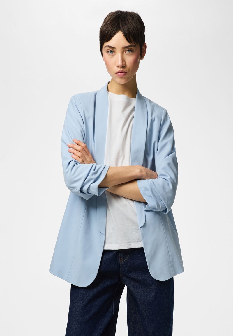 Persoon met kort donker haar staat met gekruiste armen, draagt een lichtblauwe blazer, wit overhemd en donkerblauwe broek tegen een effen achtergrond.