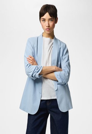 Persoon met kort donker haar staat met gekruiste armen, draagt een lichtblauwe blazer, wit overhemd en donkerblauwe broek tegen een effen achtergrond.