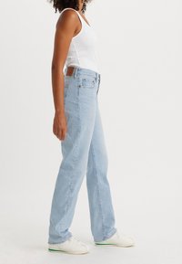 Jeans de tiro alto en azul claro, diseño de pierna recta, tela de mezclilla, con bolsillo trasero y cierre de botón, combinados con zapatos blancos y una camiseta sin mangas.