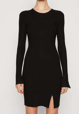 Femme portant une robe noire moulante à manches longues côtelée, avec un col rond et une petite fente à l'avant de l'ourlet.