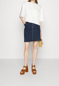 Bílý top s krátkým rukávem a volánkovými rukávy, zkombinovaný s tmavě modrou jeansovou tužkovou sukní. Hnědé platformové sandály a malá žlutá kabelka.