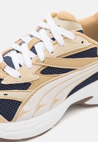 La scarpa da ginnastica beige e blu navy presenta materiali in mesh e suede, lacci bianchi e una suola in gomma testurizzata con linee di accento sui lati.