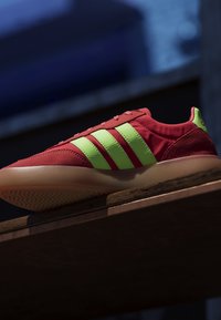 Red sneaker with three bright green stripes and light brown sole displayed on wooden surface against dark blurred background.