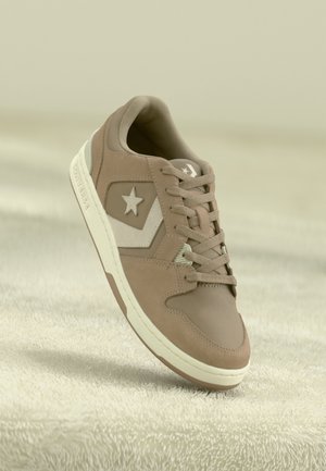 Baskets marron clair avec logo étoile et chevron blanc, semelle blanche et lacets sur une surface beige texturée.