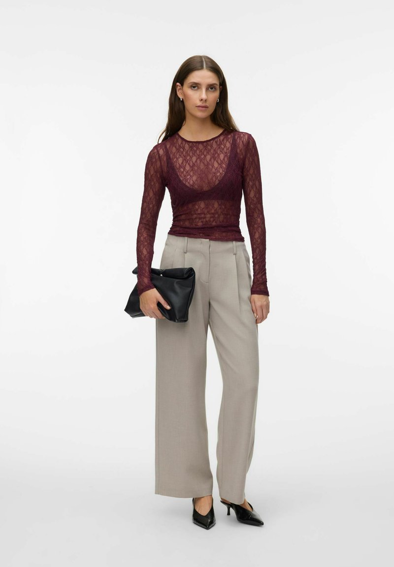 Femme portant un haut en dentelle bordeaux transparent à manches longues, un pantalon beige à jambes larges, des escarpins noirs pointus, tenant une pochette noire.
