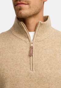 Sweater zippé beige avec un col montant, texture côtelée et détail de tirette contrastant. Le matériau semble doux et chaud.