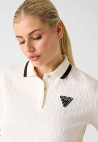 Biała koszulka polo z teksturowanym wzorem, czarnym kołnierzykiem i zatrzaskami. Z przodu czarna trójkątna naszywka z napisem "GUESS" i motywem serca.