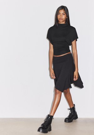 Jeune femme portant un crop top noir moulant, une jupe noire asymétrique et des bottes plateforme noires à lacets, marchant devant un mur blanc uni.