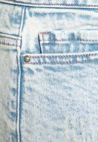 Vaaleansininen farkkukangas, jossa pieni etutasku, näkyvä oranssi ommel ja hopeinen metallinappi. Kangas on pehmeää ja siinä on haalistunut kuvio.