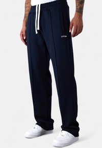 Mann in marineblauen Jogginghosen mit weißem Kordelzug und weißen Sneakern, stehend vor einem schlichten weißen Hintergrund.