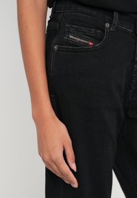 Prim plan cu partea laterală a unei persoane care poartă blugi negri Diesel, cu mâna dreaptă relaxată pe lângă coapsă.