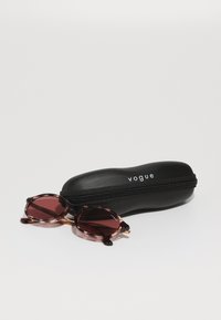 Saulesbrilles ar bruņurupuča rakstu rāmjiem un dziļi sarkaniem stikliem blakus eleganti melnai cietajai kārbai ar baltu "vogue" logo.