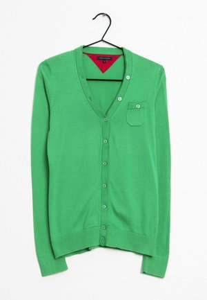 Groene gebreide cardigan met een V-hals, vijf knopen en een borstzak; gladde textuur; lange mouwen; hangend aan een zwarte haak.