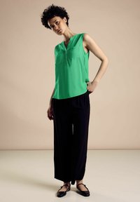 Vihreä ilmanhiha toppi V-kaula-aukolla ja kahdella etutaskulla, yhdistettynä löysiin mustiin leveälahkeisiin housuihin ja mustiin tasapohjaisiin kenkiin.