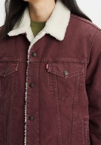 Kastanjefärgad denimjacka med krämfärgad fårskinnskrage, två bröstfickor och metallknappar. Texturerad tyg ger ett robust utseende.