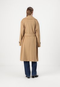 Lungo cappotto in lana di cammello con ampi lembi, vita con cintura e spalle abbassate. Indossato sopra jeans blu e scarpe nere con tacco. Design minimalista.