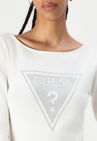 Pikkade varukatega valge kampsun, mille esiotsas on kolmnurkne hõbedaste täppidega "GUESS ?" logo. Pehme kangas laia kaelusega.