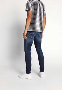 Blaue Denim-Jeans im Slim Fit mit detaillierter Naht und zwei hinteren Taschen, kombiniert mit einem schwarz-weiß gestreiften T-Shirt und weißen Sneakern.