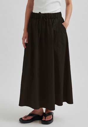 Femme portant une longue jupe noire avec taille à cordon et poches latérales, associée à des tongs plateformes noires et un haut blanc.