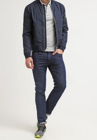 Marinblå bomberjacka med dragkedja och fickor på sidan, grå pikétröja, mörka jeans och svarta sneakers med neongrön accent.