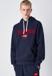 Felpa blu navy con tascone frontale, cappuccio regolabile con cordino, testo "CHAMPION" rosso in evidenza e logo accentuato sulla manica.