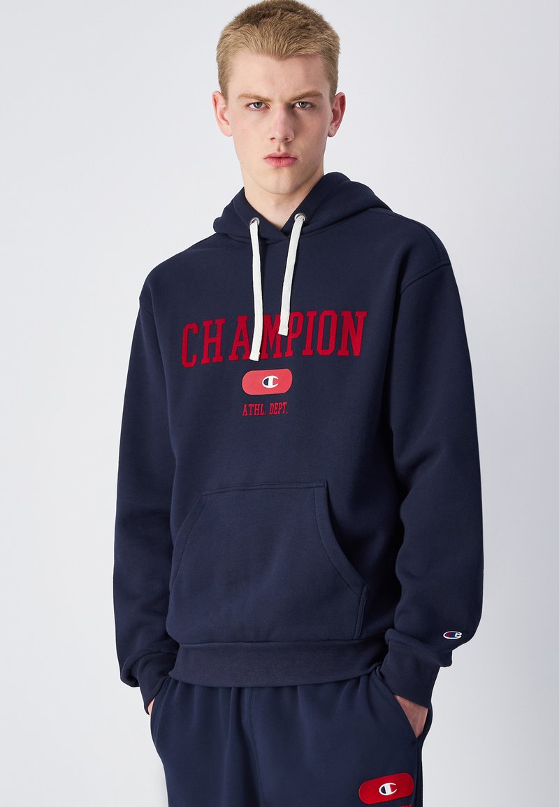 Felpa blu navy con tascone frontale, cappuccio regolabile con cordino, testo "CHAMPION" rosso in evidenza e logo accentuato sulla manica.