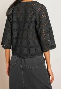 Femme portant un chemisier noir à motif œillet avec des bords en feston et une jupe en denim gris foncé, vue de dos.