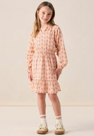 Ein langärmliges, korallenmuster Kleid mit gerafftem Taillenbereich und einem rüschengesäumten Saum. Kombiniert mit beigen Schuhen und weißen Socken mit roten Streifen.