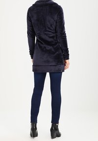 Persona in piedi di spalle, che indossa un lungo giacca plush blu scuro, jeans skinny blu scuro e stivali con tacco neri fino alla caviglia.